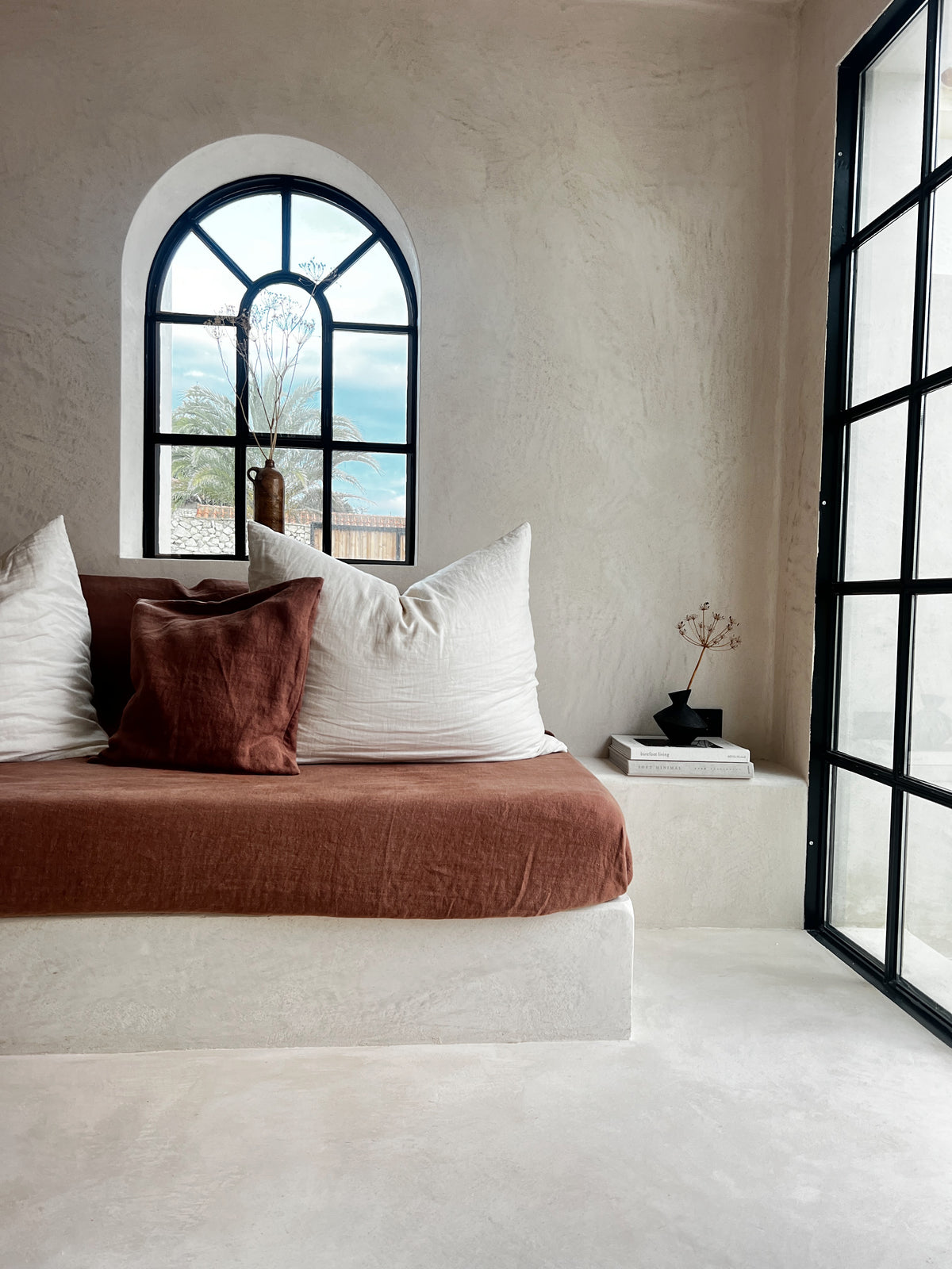 Plastered built-in daybed with terracotta cushions beside an arched window and steel-framed doors