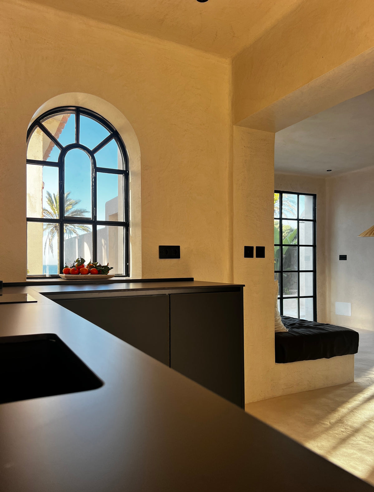 Kitchen with plastered walls, dark worktop and arched window opening onto a sitting area