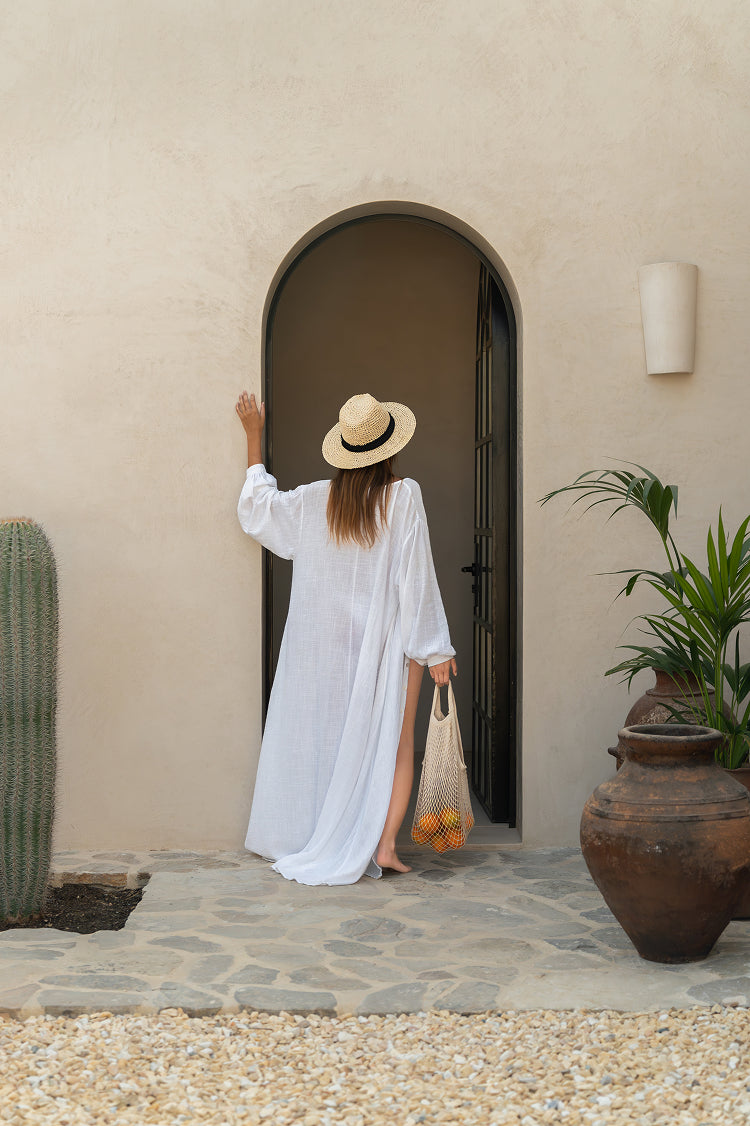 Woman in a white dress and straw hat at an arched door in a Mas Maya façade, with cacti and a clay pot