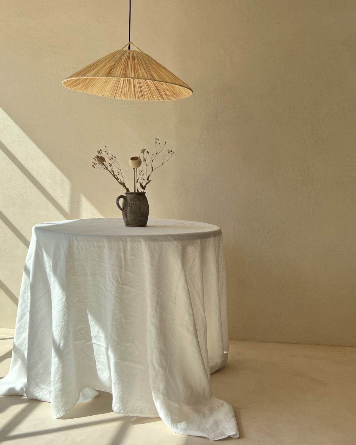 Round table with a white linen cloth and a vase of dried stems under a rattan pendant on a Mas Maya floor